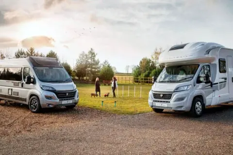Dos vehículos, un camper y una caravana, estacionados al aire libre. Dos mujeres y sus perros disfrutan de un día soleado.