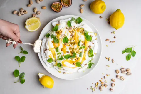 En hand skär en pavlova med lemon curd, toppad med passionsfrukt, pistagenötter och mynta.