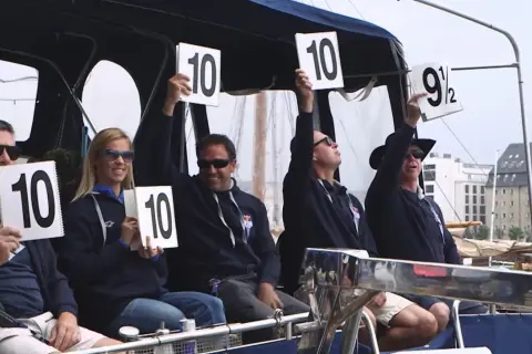 A group of diving judges hold up scorecards. Most show a perfect 10, but one judge holds up a 9.5.