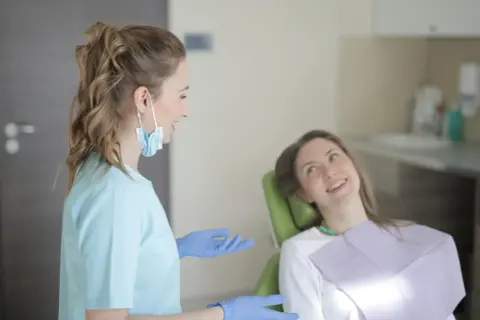 Dentystka w niebieskiej bluzie i rękawiczkach rozmawia z pacjentką. To obrazek pokazujący, jakie są studia stomatologiczne wymagania.