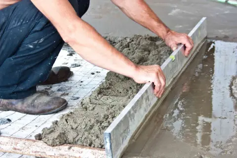 Ręce budowlańca wyrównują świeżą wylewkę betonową na zewnątrz za pomocą łaty.