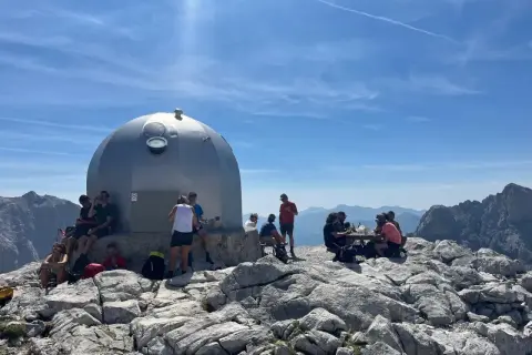 Gente descansa junto a un refugio en un viaje a Picos de Europa. El sol brilla en un cielo azul con nubes.