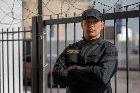 Kwalifikowany pracownik ochrony w czarnej kurtce z naszywką "SECURITY" stoi przy ogrodzeniu z drutem kolczastym.