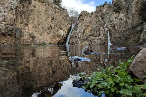 Cascadas gemelas caen entre rocas escarpadas en el embalse de Pedrezuela, reflejándose en el agua tranquila.