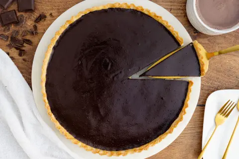 Ein Stück vom originalen Tarte au Chocolat Rezept wird mit einem goldenen Tortenheber herausgehoben. Daneben liegen Schokoladenstücke und eine Tasse Kakao.
