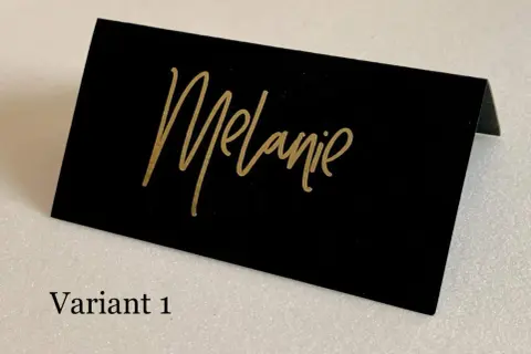 Marque place noir et or avec le prénom "Melanie" écrit en calligraphie dorée. Un élégant détail pour une table de fête.