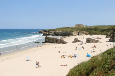 Gente disfrutando del sol y el mar en la playa Longara Barreiros, con formaciones rocosas y una casa amarilla al fondo.
