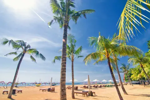 Solig strand i Sanur, Bali, med palmer, solstolar och traditionella balinesiska båtar.