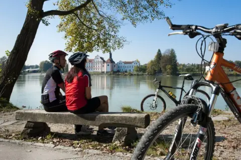 Para rowerzystów odpoczywa nad rzeką, podziwiając malownicze **trasy rowerowe Austrii** i pałac w oddali.
