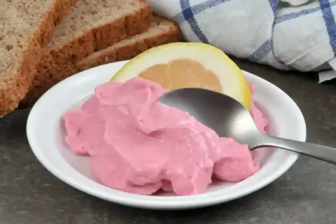 Une cuillère sert une portion de tarama rose crémeux, garnie d'une tranche de citron. Du pain grillé est visible en arrière-plan. Une recette simple pour un apéritif savoureux.