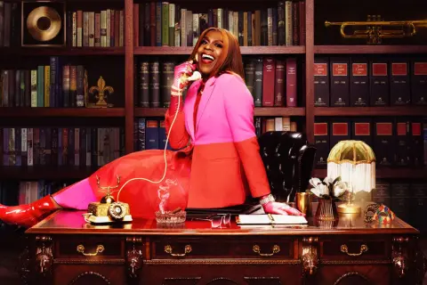 Big Freedia, in a vibrant pink and orange suit, sits at a desk, talking on a vintage phone. Her success is evident in the opulent office, hinting at her impressive big Freedia net worth.