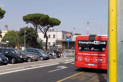 Czerwony autobus Ciampino Airlink czeka na pasażerów. Z tyłu widnieje napis "FROM TERMINAL TO ROME TERMINI" i cena 2,70€. W tle parking z samochodami i budynek lotniska.