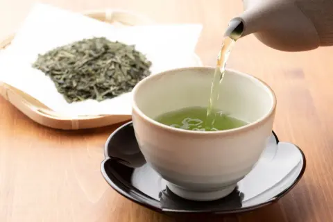 Comment faire du thé vert : une théière verse un liquide vert vif dans une tasse, à côté d'une pile de feuilles de thé séchées.