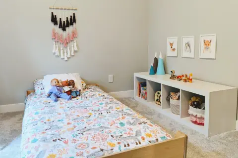 A cozy floor bed setup with a woodland-themed duvet cover, dolls, and a wall hanging. A white cube shelf holds books and toys.