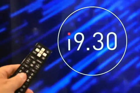 Dłoń z pilotem przed ekranem z godziną 19.30. Czy zmiana godziny emisji programu wpłynęła na oglądalność?