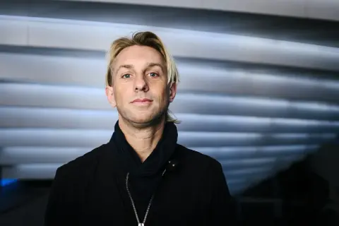 Anders Hansen, din personlighet speglas i blicken. En man med karisma, klädd i svart jacka och scarf, mot en bakgrund av horisontella linjer.