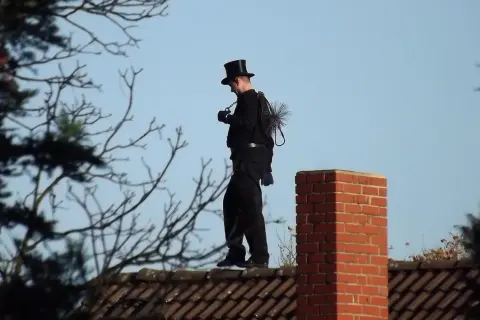 Ein Schornsteinfeger in Zylinder und schwarzer Kleidung steht auf einem Dach. Er trägt seine Bürste und Kette bei sich. Man fragt sich, wie oft kommt der Schornsteinfeger wohl?