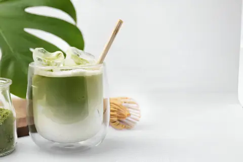Un verre de thé matcha glacé, parfait pour savoir si on peut boire le thé matcha froid. Une feuille de monstera et un fouet à matcha complètent la scène.