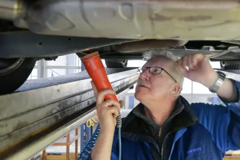 Mechaniker prüft Auto auf Hebebühne mit Lampe. Hoffentlich hat er den Fahrzeugschein dabei, sonst gibt's Ärger!