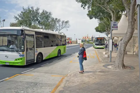Kobieta czeka na autobus na przystanku przy lotnisku na Malcie. W tle widać inne autobusy i budynki.