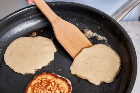 Buttermilch pancakes braten in der Pfanne. Ein Holzspatel wendet einen goldbraunen Pancake.