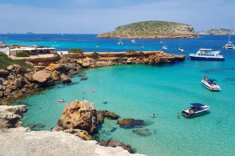 Plage idyllique à Ibiza avec eaux turquoise, rochers, bateaux et baigneurs. Parfait pour savoir où aller à Ibiza.