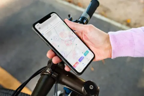 Dłoń trzyma smartfon z mapą i ikonami aplikacji rowerowych, oparty o kierownicę roweru.