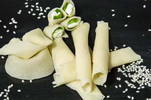 Rotolini di crepes con farina di riso, ripieni di formaggio fresco e basilico, sparsi su un tavolo scuro con semi di sesamo.