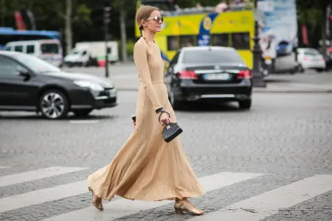 Elegancka, beżowa sukienka maxi, idealna na miejskie stylizacje. Kobieta w okularach przeciwsłonecznych i małej czarnej torebce przechodzi przez pasy.