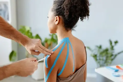Therapeut klebt blaues Tape auf die Schulter einer Frau, um **Schulterschmerzen im Ruhezustand** zu lindern.