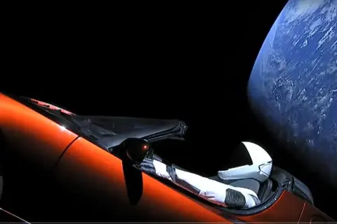 Pomarańczowy Tesla Roadster z manekinem w skafandrze kosmicznym dryfuje w kosmosie obok Ziemi.