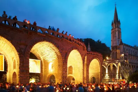Eine Lichterprozession in Lourdes. Menschen halten Kerzen unter einem steinernen Bogen, während im Hintergrund eine Kirche beleuchtet ist.