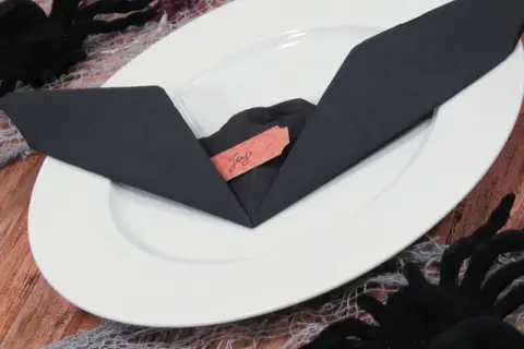 Une serviette noire pliée en forme de chauve-souris sur une assiette blanche, avec une étiquette "Joy". Décoration d'Halloween.
