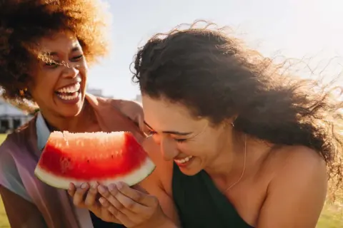 Zwei lachende Frauen teilen ein Stück Wassermelone. Perfekt für einen heißen Tag, da Wassermelone kalorienarm ist und erfrischt.