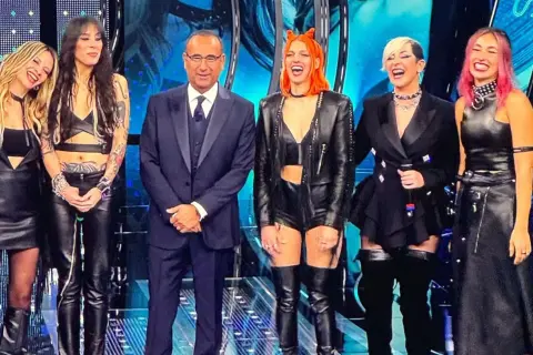 Micky Paiano con un completo scuro, affiancato da quattro donne con outfit in pelle audaci e colorati, sorridono sul palco.