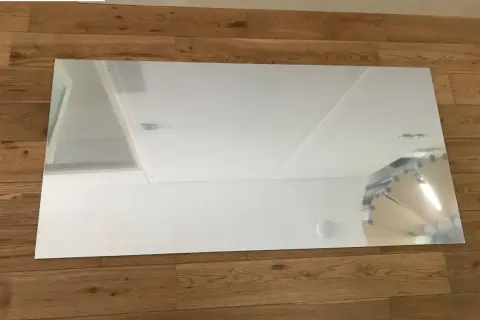 Un grand panneau de dibond miroir reflète le plafond et un mur blanc, posé sur un parquet en bois.