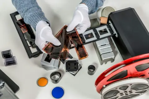 Händer med vita handskar sorterar filmremsor och diabilder för att omvandla negativ till digitala bilder. En filmscanner och en filmrulle syns också.