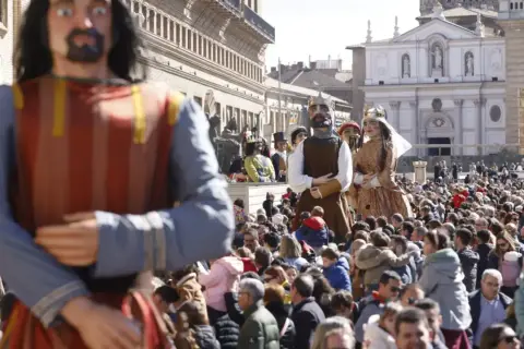 Gigantes y cabezudos desfilan entre una multitud en una plaza. Las figuras de cartón piedra, algunas con coronas, son el centro de atención.