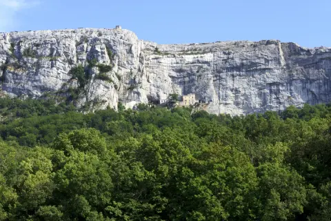 Vue du pic de Bertagne, une falaise imposante surplombant une forêt verdoyante. Un petit bâtiment est visible sur le flanc de la falaise.