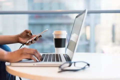 Kobieta pracuje na laptopie, trzymając telefon. Obok stoi kubek kawy. To idealne miejsce do planowania strategii rozwoju firmy, być może z uwzględnieniem wsparcia pomoc de minimis.