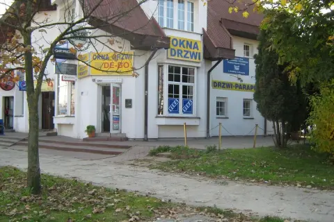 Budynek z żółtymi banerami "OKNA DRZWI" i "DRZWI PARAPETY", z drzewem po lewej.