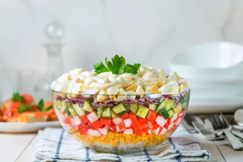 Deliciosa ensalada de surimi en capas, con maíz, pepino, cebolla morada y huevo duro, cubierta de mayonesa y perejil.