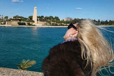 Kobieta z blond włosami podziwia pomnik w Brindisi. W tle błękitne morze i błękitne niebo.