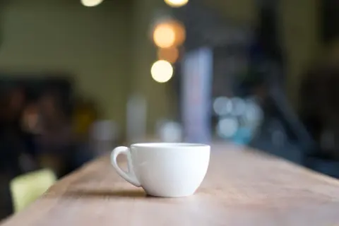 En vit, tom mått kaffekopp står på ett träbord. Bakgrunden är suddig med ljus.