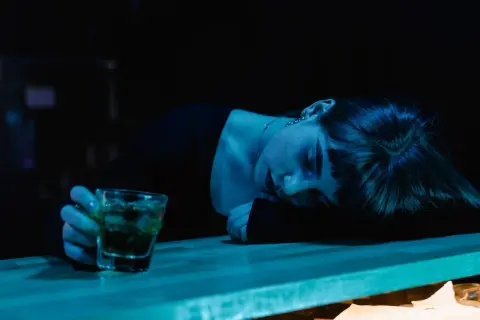 Zmęczona kobieta opiera głowę na blacie, obok szklanka z drinkiem. Mąż alkoholik obwinia mnie o wszystko, czuję się wyczerpana.