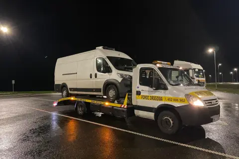 Nocne holowanie na drodze ekspresowej. Biała furgonetka jest transportowana na lawecie pomocy drogowej.