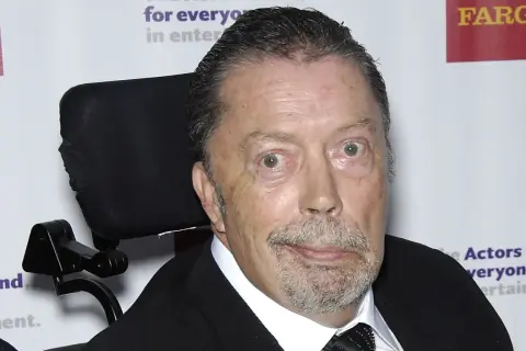 Tim Curry w wózku inwalidzkim, zmagający się z chorobą, ale wciąż z iskrą w oku.