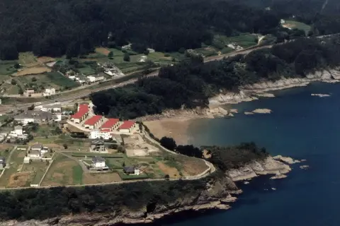 Vista aérea de casas con tejados rojos cerca de la playa de Sacido, rodeada de vegetación y el mar azul.