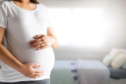 Kobieta w białej koszulce trzyma dłonie na zaokrąglonym brzuchu ciążowym.