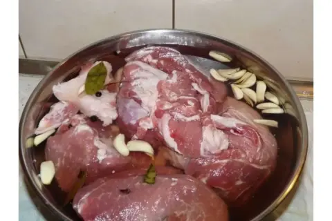 Mięso w misce, przygotowane do peklowania kurczaka na mokro do wędzenia. Widać kawałki mięsa, czosnek, liście laurowe i ziarna pieprzu.